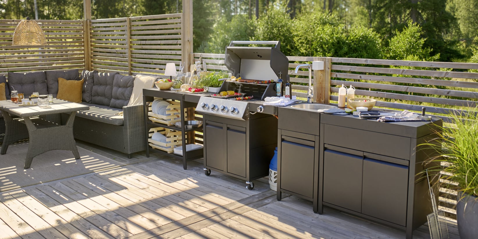 En uteplats med en flätingssoffa och ett soffbord nära en grill och förberedelsestation med mat. Träpaneler och växter pryder bakgrunden.