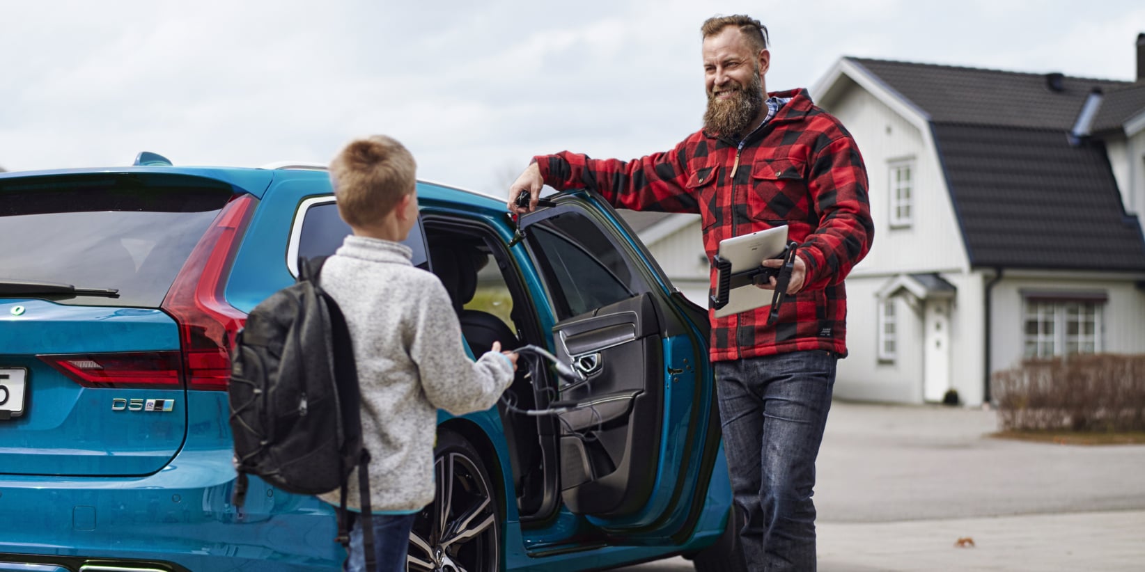 En man i en rödrutig jacka vid en öppen bildörr ler mot ett barn med ryggsäck. Hus kan ses i bakgrunden.