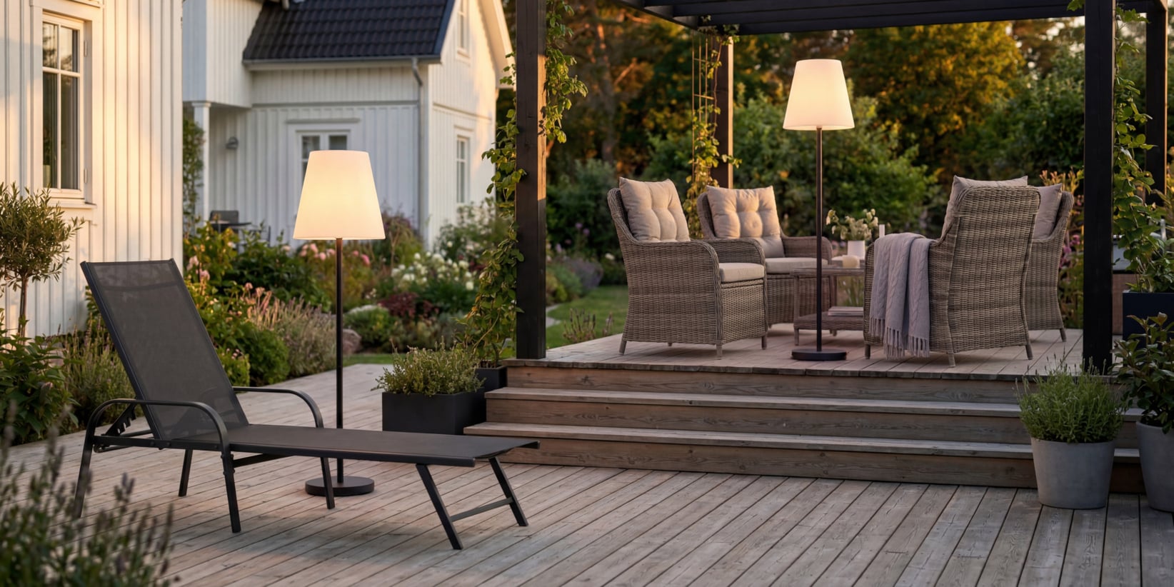 Weiden-Möbel, ein Esstisch, Stehlampen und Pflanzen stehen auf einer hölzernen Terrasse mit Stufen bei einem weißen Haus mit Blick auf einen Garten.