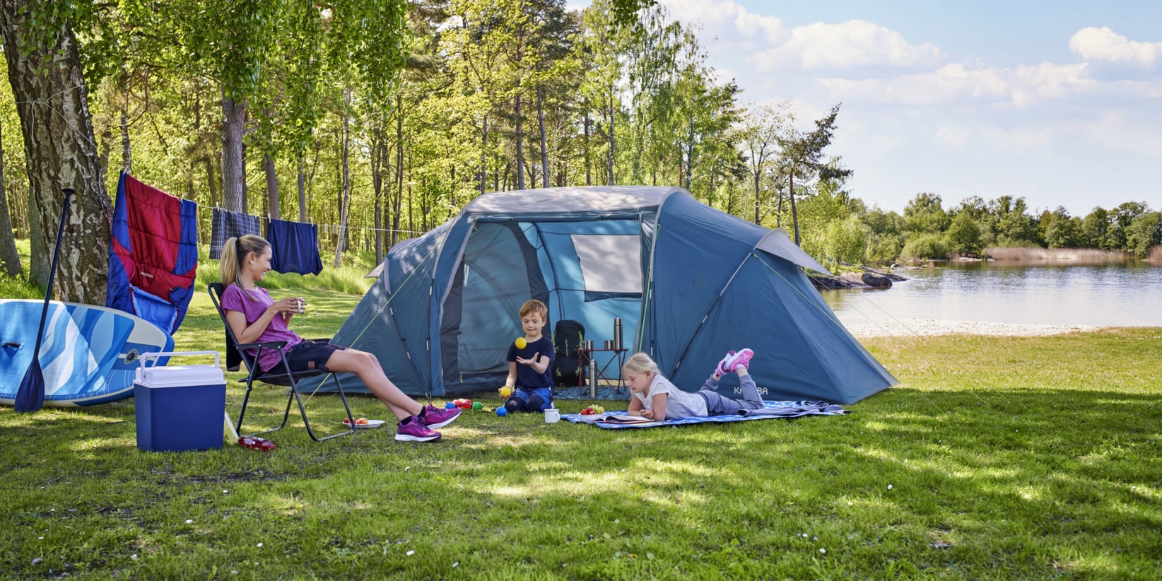 En person slappnar av i en campingstol vid ett blått tält, medan två barn leker på en filt i gräsbevuxen skog vid en sjö, med tvätt som torkar i närheten.