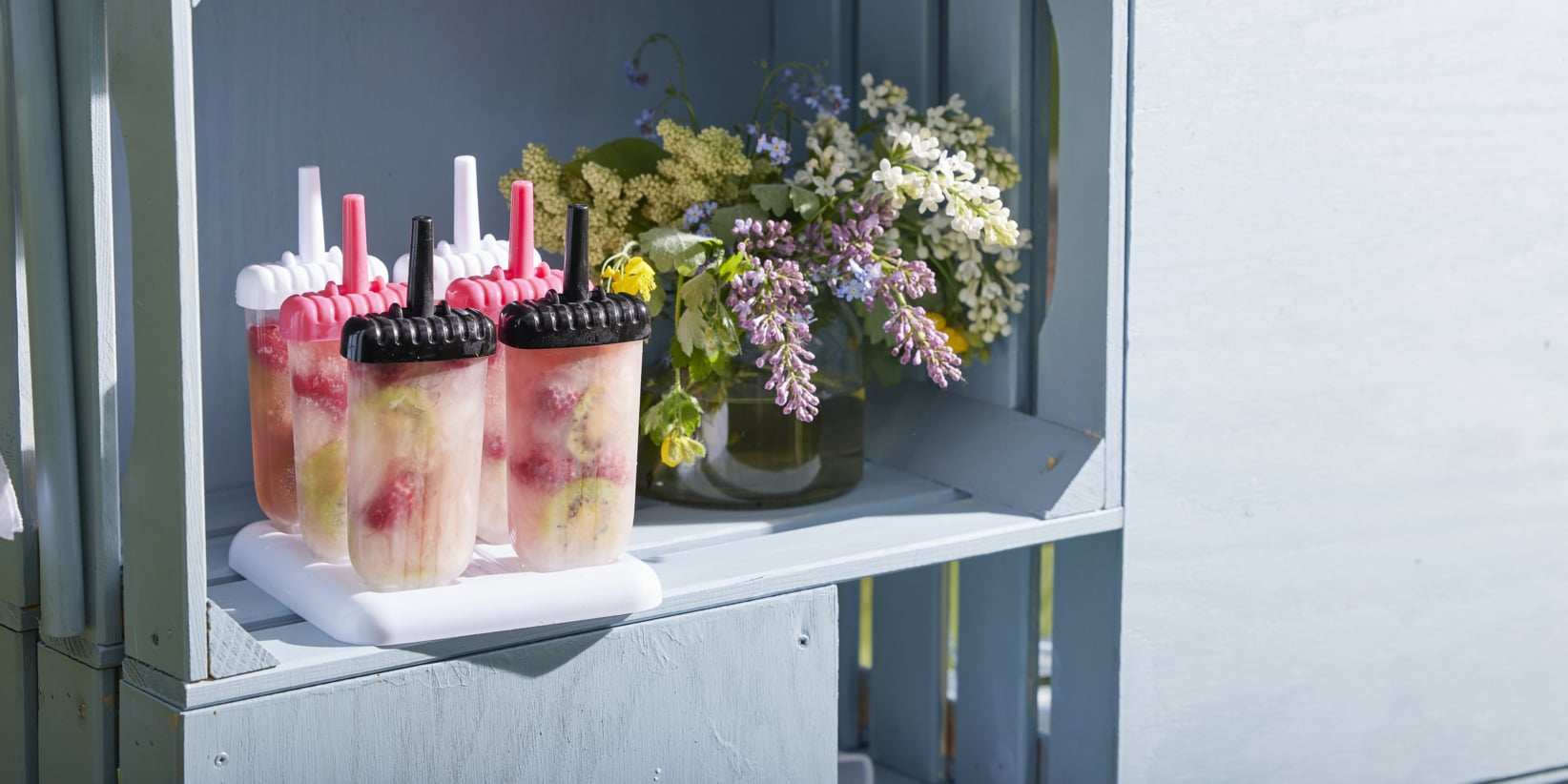 Fyra fruktisglass med bär och kiwi i klara formar står i en vit hållare på en ljusblå hylla, med vilda blommor bakom.