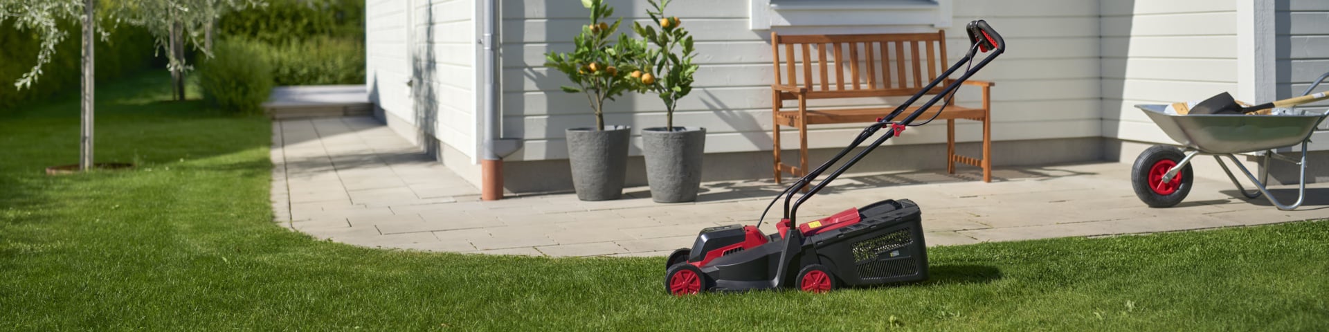 Ein schwarz-roter Rasenmäher steht auf Gras in der Nähe eines Hauses, mit Topfpflanzen, einer Holzbank und einer Schubkarre auf der Terrasse.