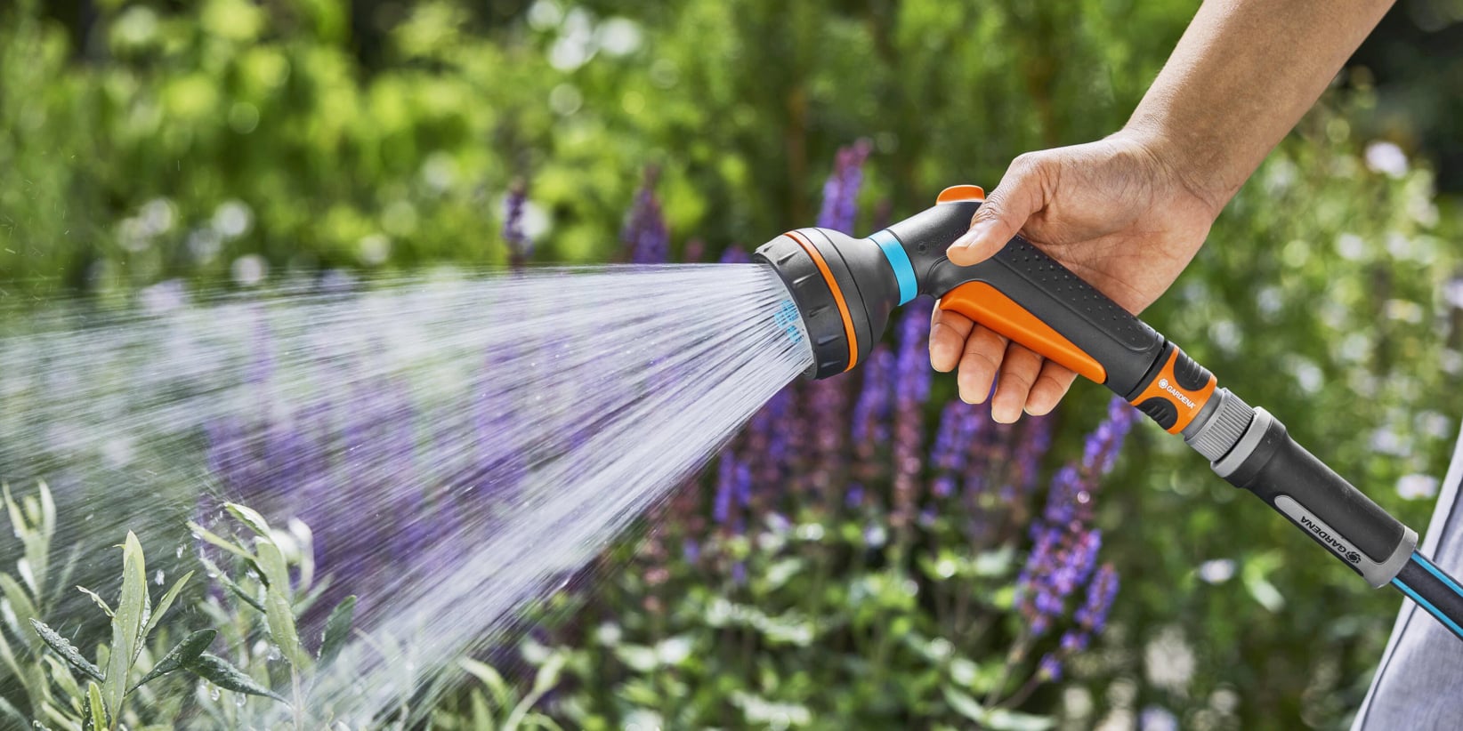 Eine Hand hält eine orange-schwarze Schlauchdüse, gießt üppig grüne Pflanzen und blühende violette Blumen in einem Garten.