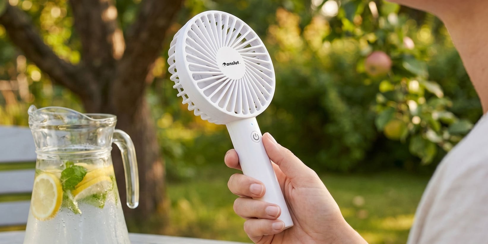 Jemand benutzt draußen einen weißen Handventilator, mit einem Krug Zitronen-Minzwasser auf dem Tisch und laubigen Bäumen im Hintergrund.