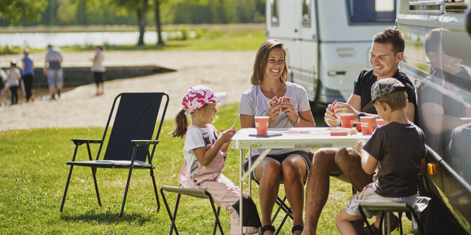 Fyra familjemedlemmar spelar kort vid ett utomhusbord vid en lägerdeltagare. Två tomma stolar, grönt gräs, träd och människor vid vattnet är synliga.