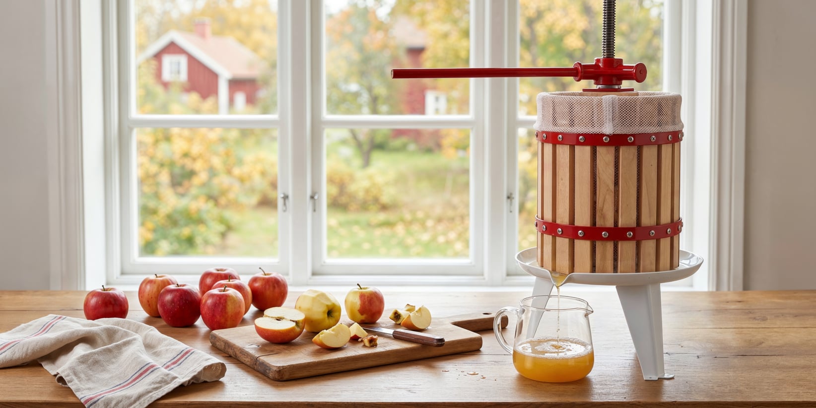 En trääppelpress pressar juice i en glaskanna på ett bord bredvid ett fönster. Runt den finns hela och skivade äpplen, en kniv och ett randigt tyg, med ett höstlandskap synligt genom fönstret.
