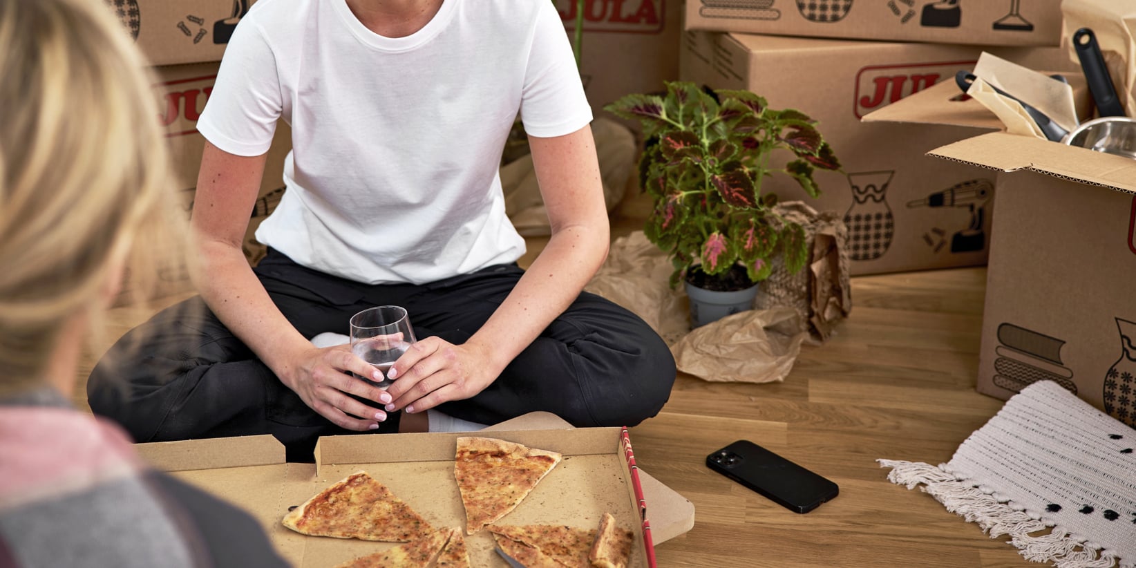 En person i en vit t-shirt sitter med benen i kors på golvet bredvid en öppen pizzakartong och ett glas vatten. Bakom dem står kartonger, en växt, en telefon och olika oupppackade tillhörigheter, vilket visar att de just har flyttat in.