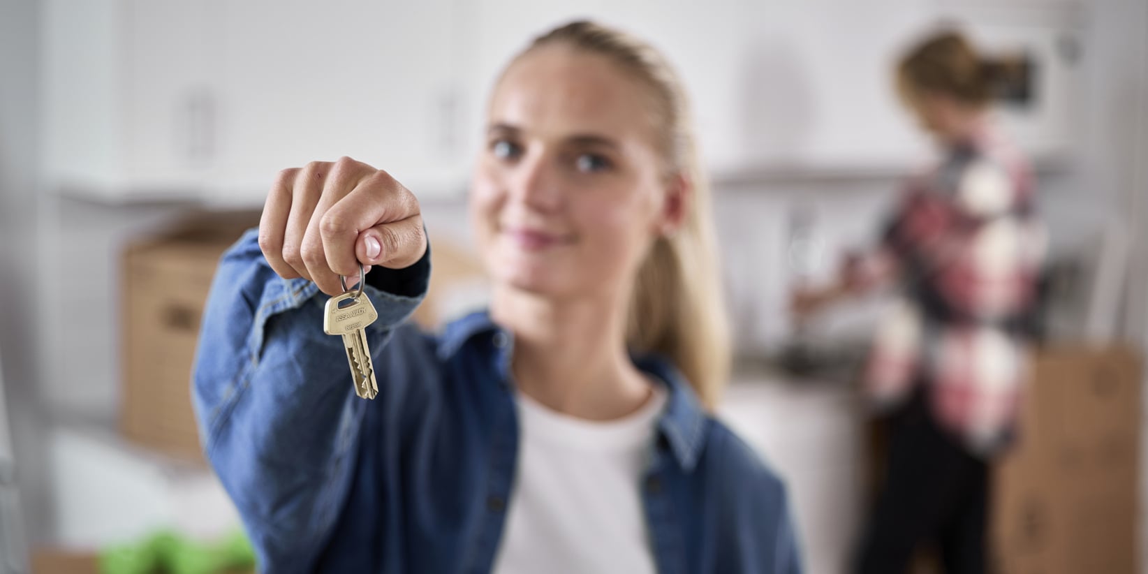 En nyckelknippa hålls framträdande fram av en person, medan en annan packar upp lådor i ett kök bakom dem. Hemmiljön tyder på att de kanske flyttar in eller bosätter sig i en ny bostad.