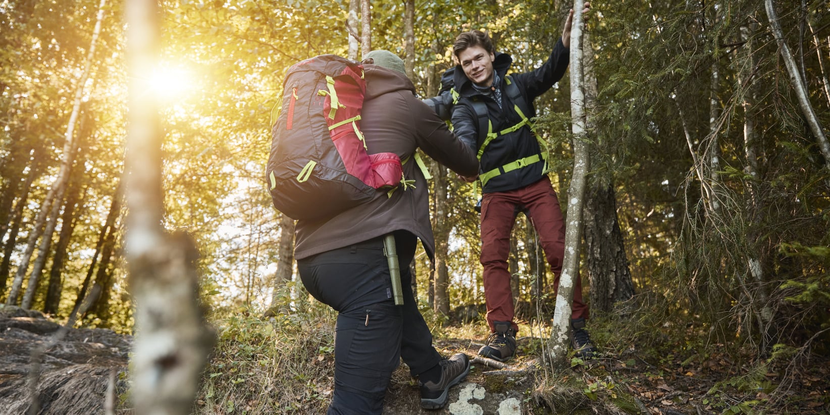 En person hjälper en annan uppför en skogsklädd sluttning. Båda är utrustade med ryggsäckar och klädda för utomhusmiljö. Solljuset strömmar genom träden och lyser upp en ojämn mark täckt av stenar, grenar och utspridda löv.