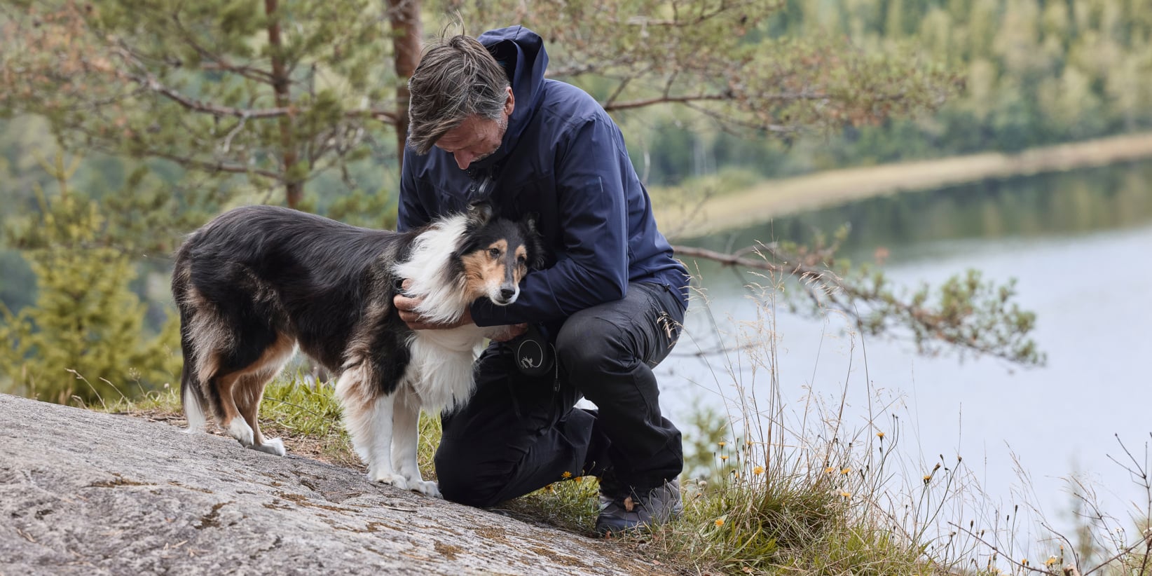 En person i marinblå jacka knäböjer bredvid en svart, vit och beige hund på stenig mark vid en sjö, med gräsytor och träd i bakgrunden.