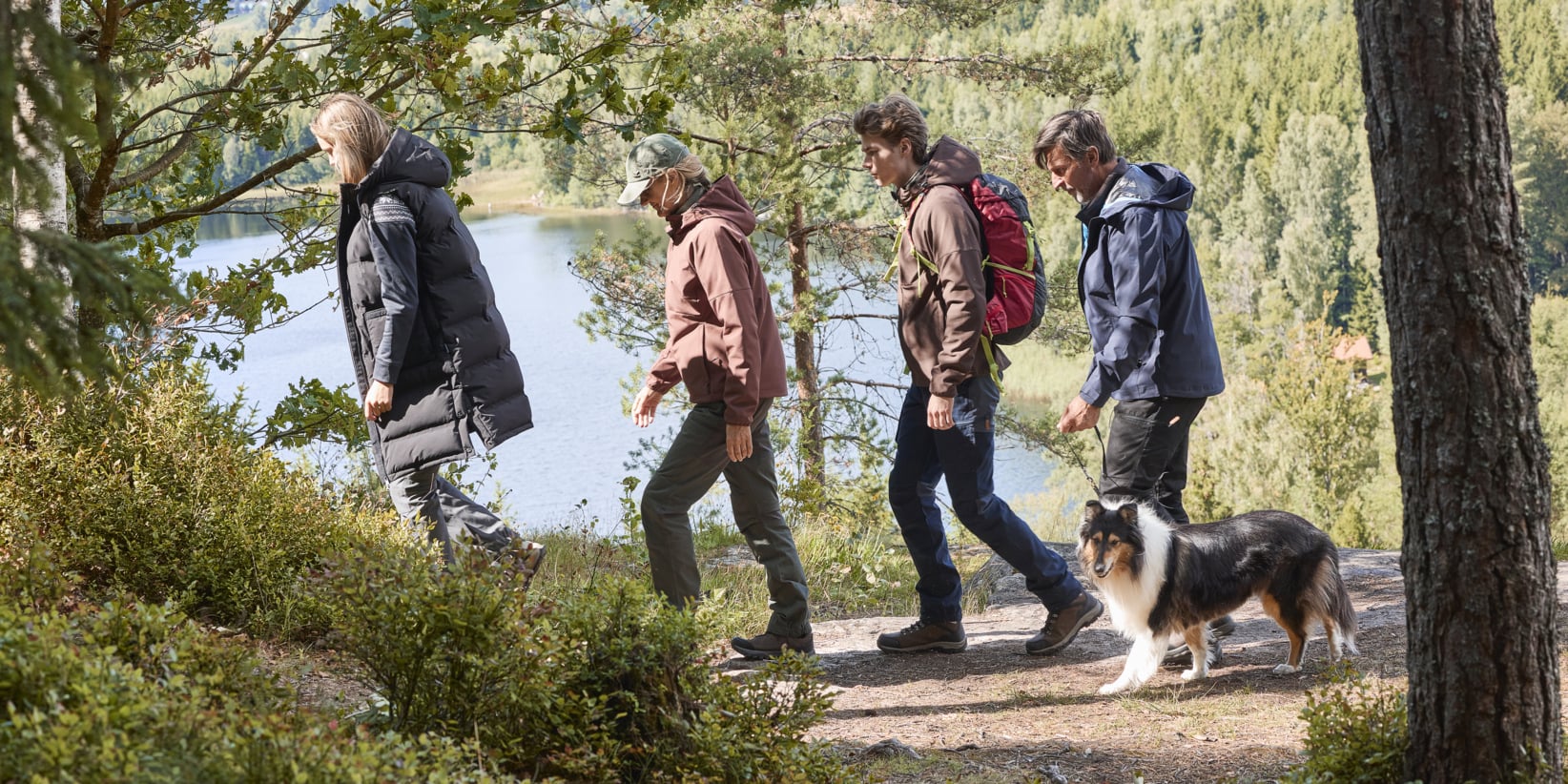 Fyra personer och en hund vandrar längs en skogsstig intill en sjö, omgivna av träd och frodig grönska. Gruppen bär utomhuskläder, med sjön synlig bakom dem.
