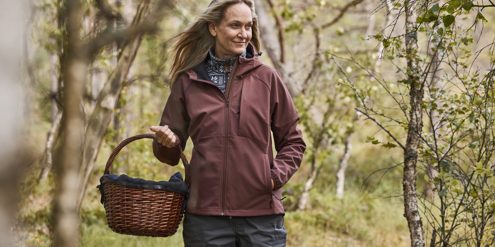 Eine blonde Frau in einer kastanienbraunen Jacke läuft durch einen laubreichen Wald, hält einen Weidenkorb in der linken Hand und blickt zur Seite. Üppig grüne Bäume umgeben sie.