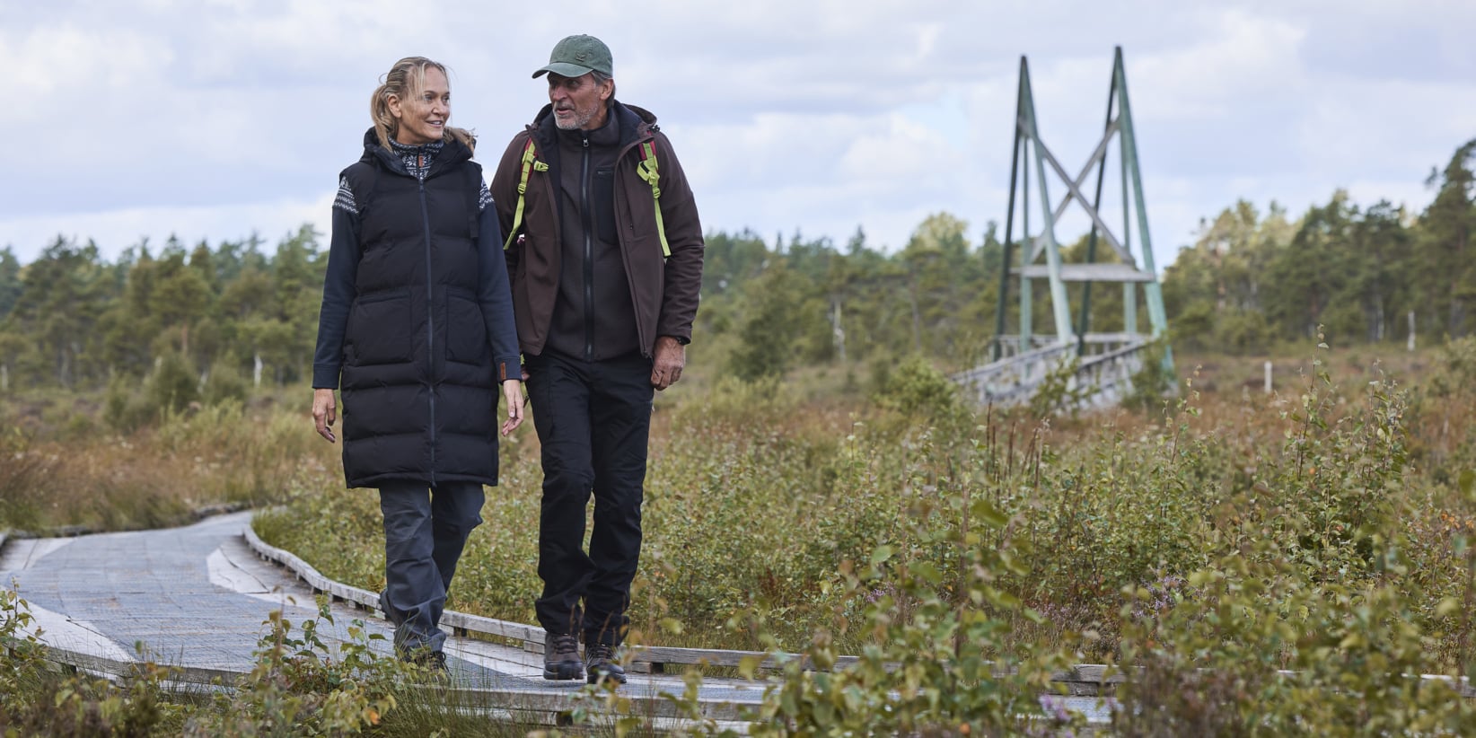 Två personer promenerar bredvid varandra längs en trästig omgiven av frodig grönska, med en bro i fjärran. De bär svalväderskläder för utomhus.