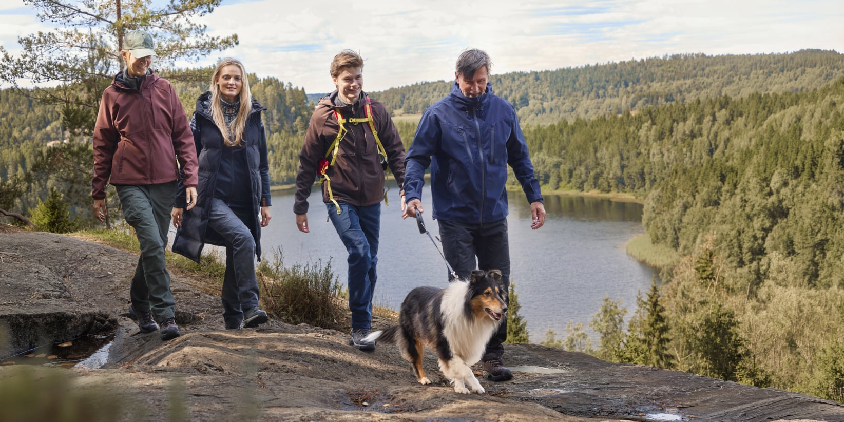 Vier Personen und ein Hund wandern auf einem felsigen Pfad mit Blick auf einen See und baumbewachsene Hügel unter einem wolkenübersäten Himmel.
