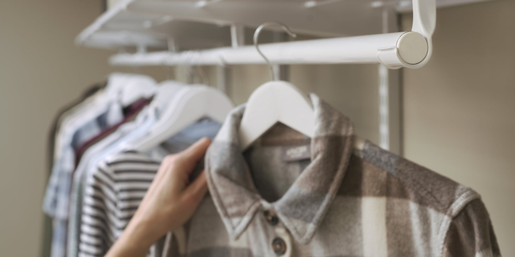 Käsi tarttuu ruudulliseen paitaan, joka roikkuu valkoisessa henkarissa erilaisten paitojen joukossa, mukaan lukien raidallisia kuvioita, jotka on esillä vaatetelineessä.