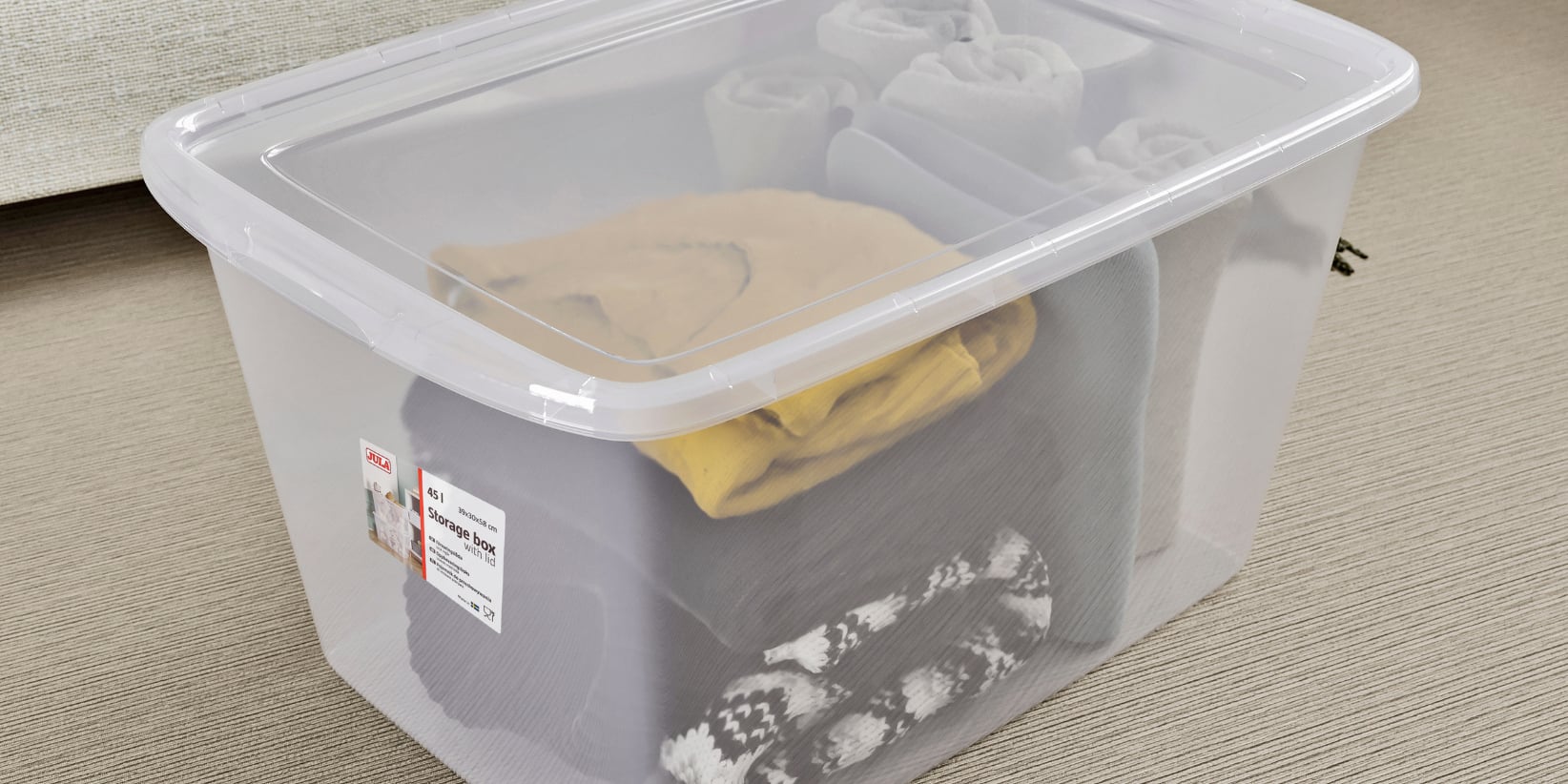 En gjennomsiktig plastboks med lokk inneholder pent brettede klær, for eksempel en gul skjorte, noen grå plagg og et plagg med svart-hvitt mønster. Fire sammenrullede plagg ligger bakerst i boksen.