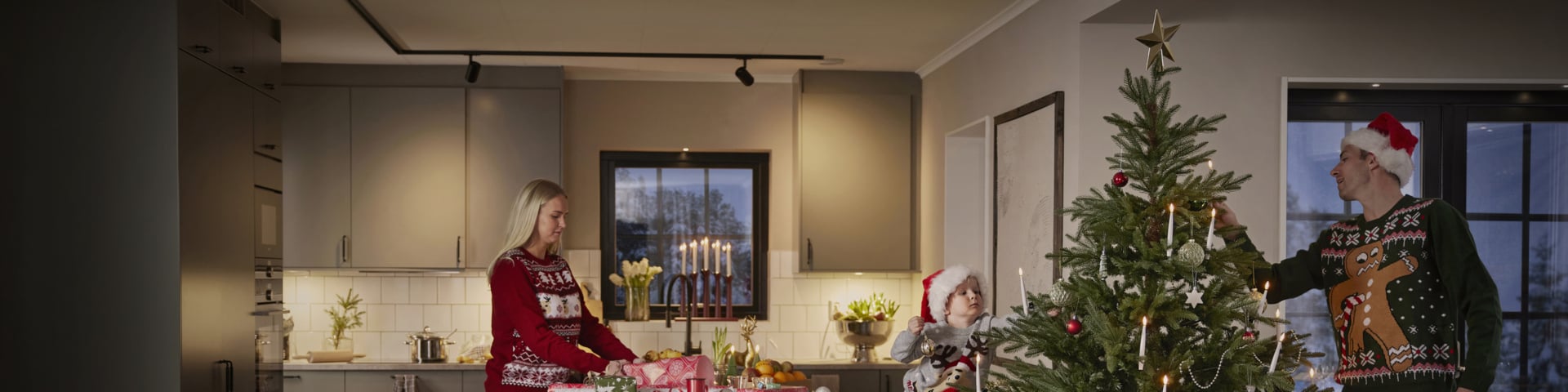 Eine Frau in einem festlichen Pullover packt Geschenke an der Küchentheke, während ein Kind und ein Mann in Weihnachtspullovern und Weihnachtsmützen gemeinsam einen Weihnachtsbaum in ihrem modernen Zuhause schmücken.
