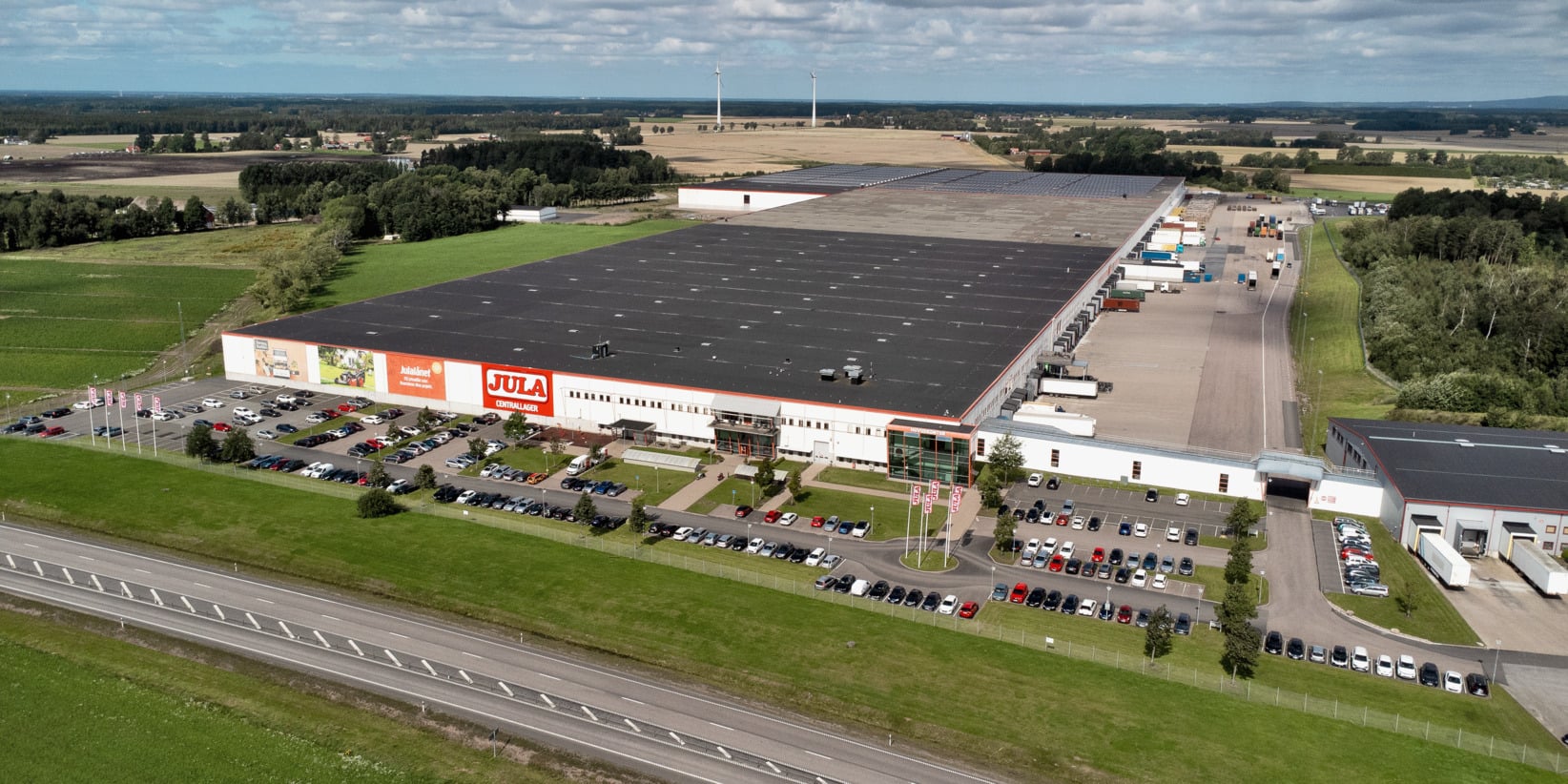 Ett stort lager med en "Jula"-logotyp på framsidan, omgivet av en parkeringsplats full av bilar med närliggande gräsmattor, vägar och fält. Flera lastbilar står stationerade vid lastkajer längs sidan.