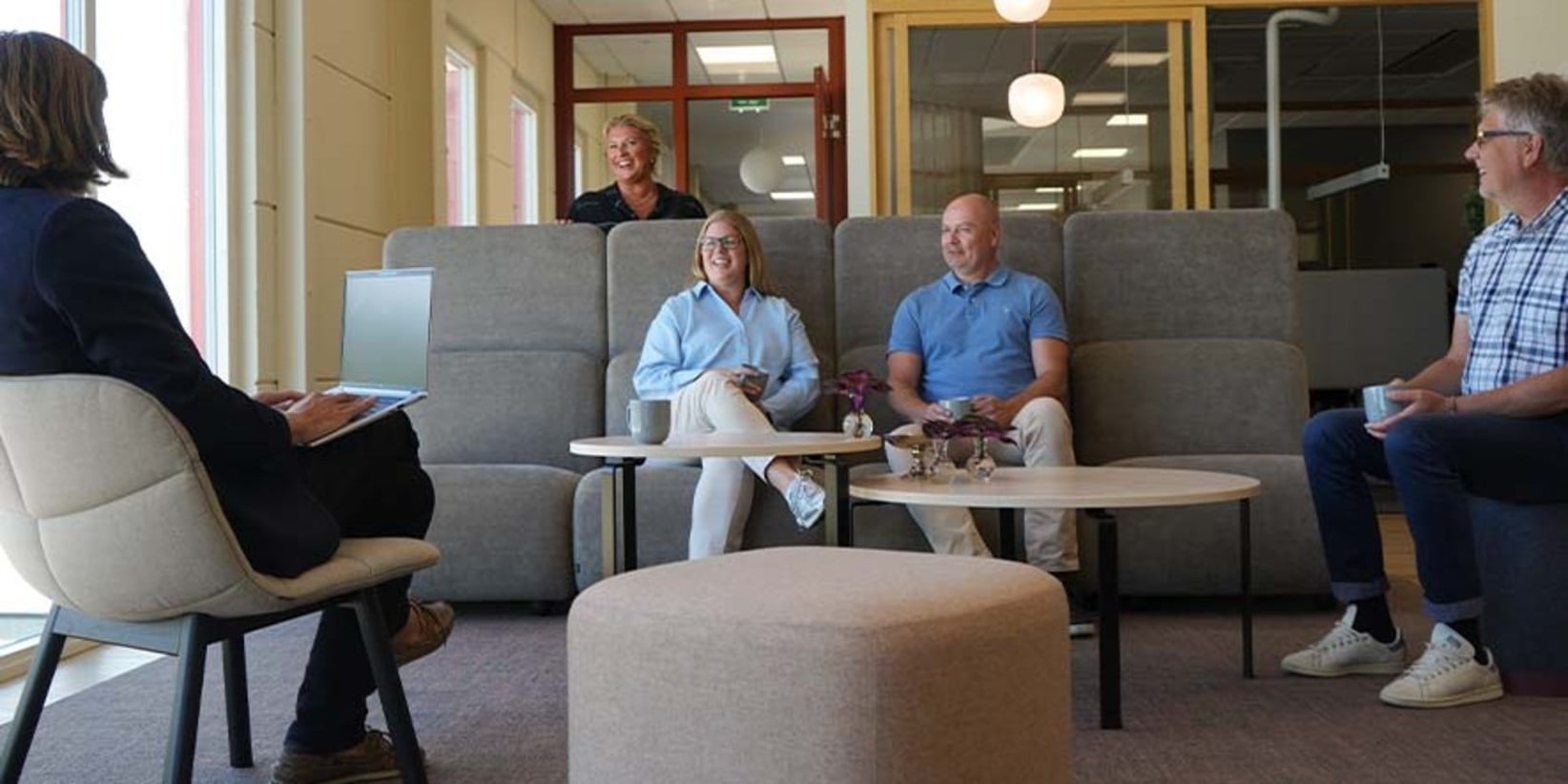 Fem vuxna har ett informellt möte i en lounge, några sittande och andra stående runt beige stolar och bord. En laptop, koppar och en växt kan ses.