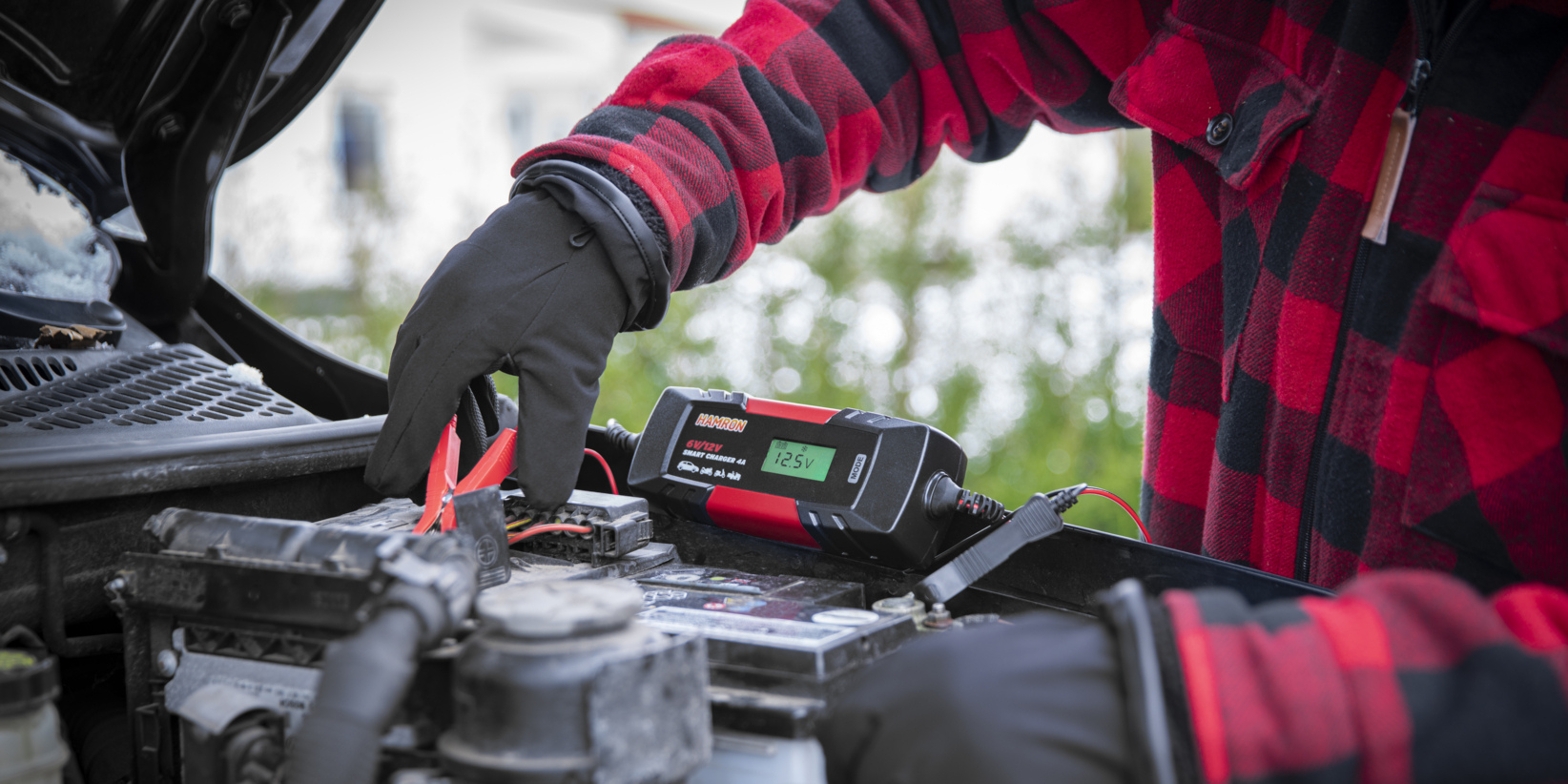 Eine Person, die schwarze Handschuhe und ein rotkariertes Hemd trägt, bedient ein Batterieladegerät an einem Automotor, wobei der Gerätebildschirm 12,5 V anzeigt, da sie mit den Batterieanschlüssen unter der offenen Motorhaube verbunden ist.