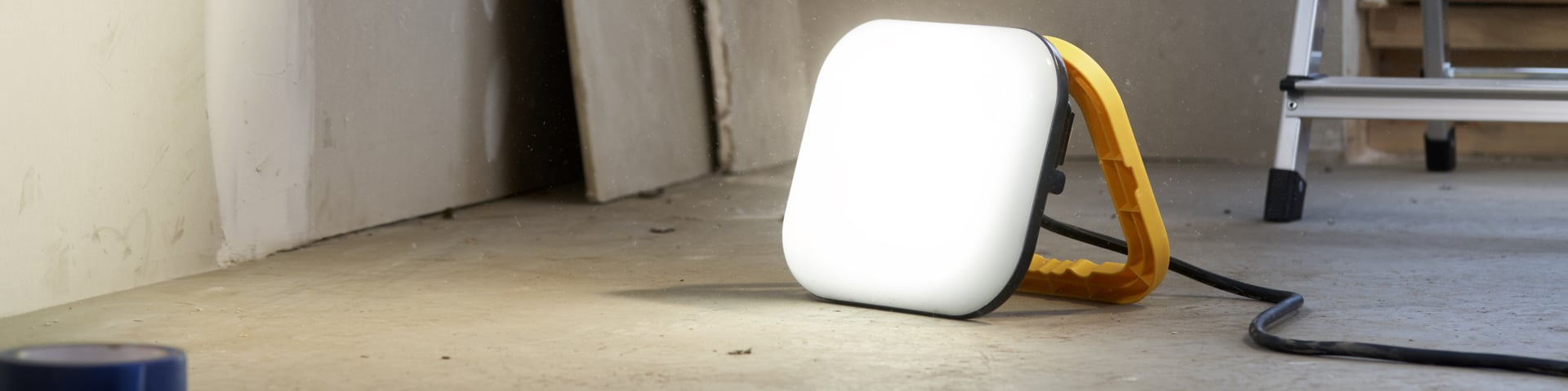Tragbare LED-Arbeitslampe mit einem quadratischen, beleuchteten Panel und gelbem Griff, der auf einem Betonboden steht.