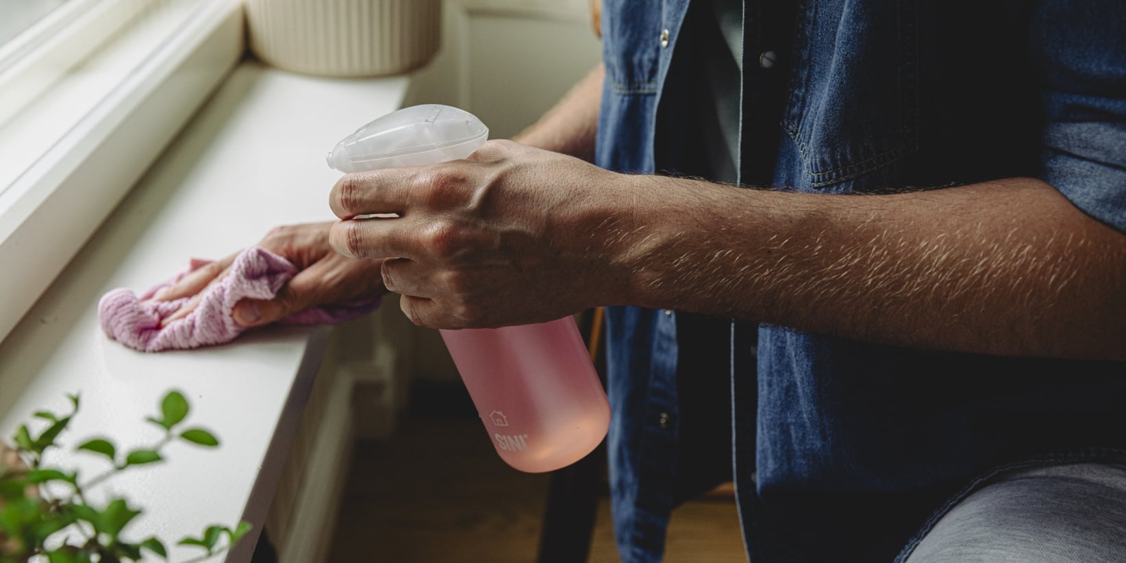 En person som håller i en rosa sprayflaska torkar av en fönsterbräda med en rosa trasa.