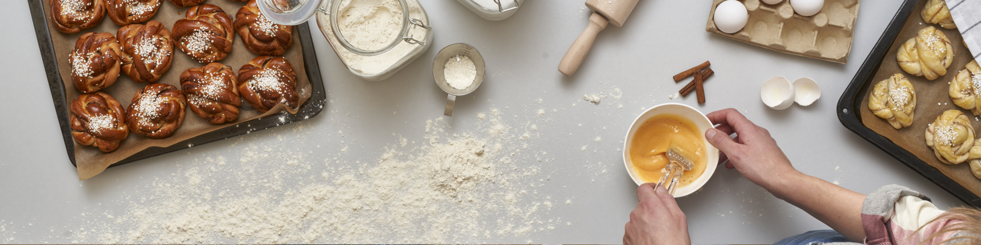 Eine Person verquirlt Eier in einer Schüssel, die von Backzutaten umgeben ist, darunter ein Blech mit gebackenen Zimtschnecken, ein Glas Mehl, Zimtstangen und ein Blech mit ungebackenen Brötchen.