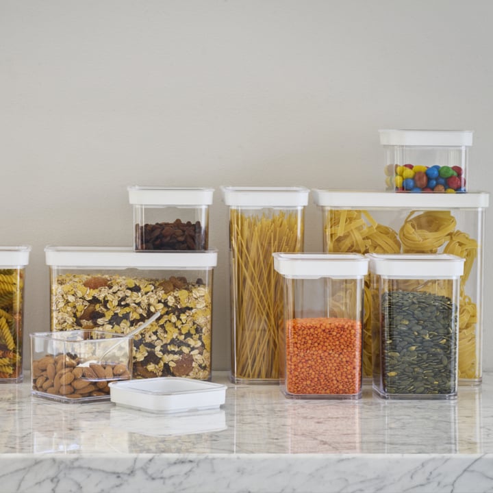 En mängd genomskinliga plastbehållare med vita lock står uppställda på en bänkskiva av marmor. I behållarna finns torrfoder som pasta, linser, havre, mandel, russin, pumpakärnor och färgglatt godis.