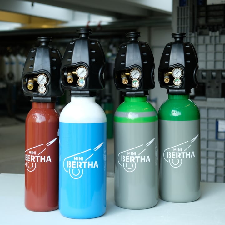 Fyra gasflaskor märkta Mini Bertha, var och en i olika färger (röd, blå, grå och grön), står upprätt på ett bord inomhus, utrustade med svarta regulatorer och mätare på toppen.