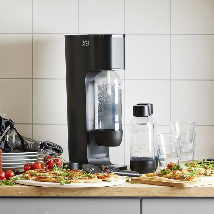 En svart AGA läskmaskin med en kolsyreflaska står på en köksbänk bredvid en mindre flaska, två glas, tallrikar med pizza med grönsaker, körsbärstomater och staplade tallrikar mot en kaklad vägg.