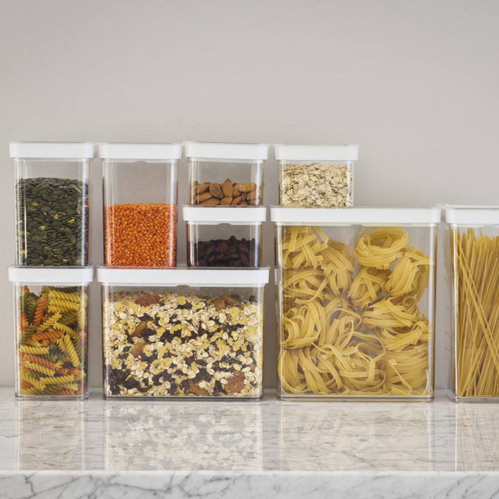 På en benkeplate av marmor står det klare plastbeholdere med hvite lokk, fylt med tørrmat som gresskarkjerner, røde linser, mandler, rosiner, havregryn, spiralpasta, müsli, tagliatelle-reir og spaghetti.