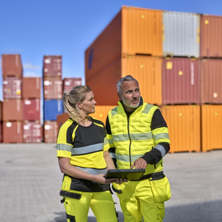 To arbeidere i advarselsklær står på et skipsverft og holder et brett i hendene. I bakgrunnen ser vi stablede containere og en kran, og himmelen er blå.