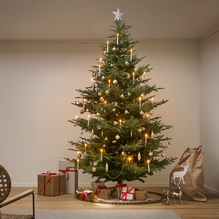 Et juletre med hvite lys, gull og hvit pynt, og en stjerne på toppen står i en stue med gaver under.