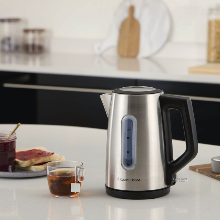 En elektrisk vannkoker i rustfritt stål på en benk ved siden av te i et glass, syltetøy og toast. Et moderne kjøkken med skap ligger bak.
