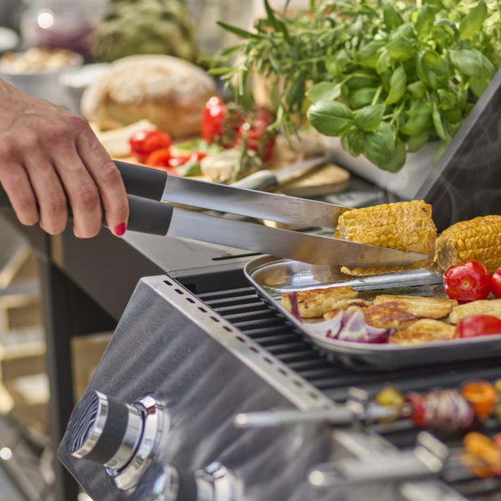 Za pomocą szczypiec dodaje się kukurydzę na kolbie na tacę z grillowanymi warzywami na grillu; W tle stoją świeże zioła i chleb.