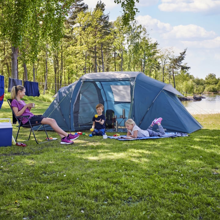 Eine Frau entspannt sich in einem Campingstuhl neben einem blauen Zelt, während zwei Kinder auf einer Decke spielen; Bäume, ein See und eine Wäscheleine liegen hinter ihnen.