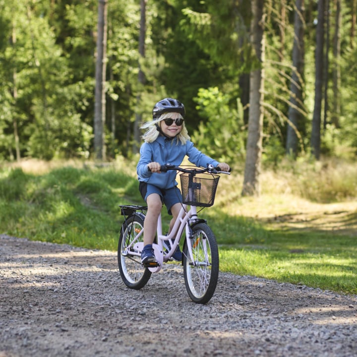 Ein behelmtes Kind in Blau, mit Sonnenbrille, fährt auf einem pinken Fahrrad mit einem Korb einen Schotterweg zwischen üppig grünen Bäumen.