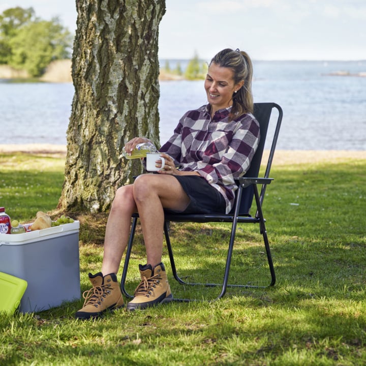 Eine Person auf einem schwarzen Klappstuhl gießt Wasser in eine Tasse am See, mit einer Kühlbox, Essen, Stiefeln, Bäumen und Wasser an einem sonnigen Tag.