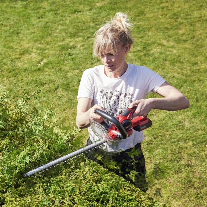 Eine Person schneidet grüne Büsche in einem grasbewachsenen Garten mit einem rot-schwarzen kabellosen Heckenscher, trägt ein weißes T-Shirt mit Buchstaben und gebundene Haare.