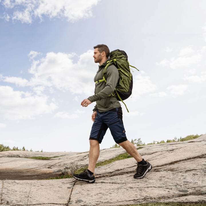 En person i en grön hoodie, blå shorts och en stor grön ryggsäck går över stenig terräng under en delvis molnig himmel.