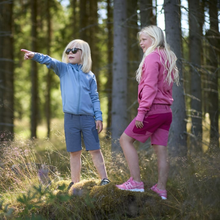 Två blonda barn står på en mossig sten i en skog; En i blått pekar framåt medan den andra i rosa ser ut så också.