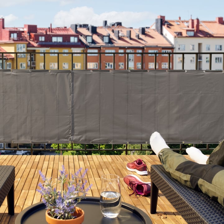 Jemand liegt auf einem Balkon mit Sichtschutzgitter, Holzboden, Liegestühlen, einem Tisch mit Getränken, Topfpflanze und Gebäuden dahinter.