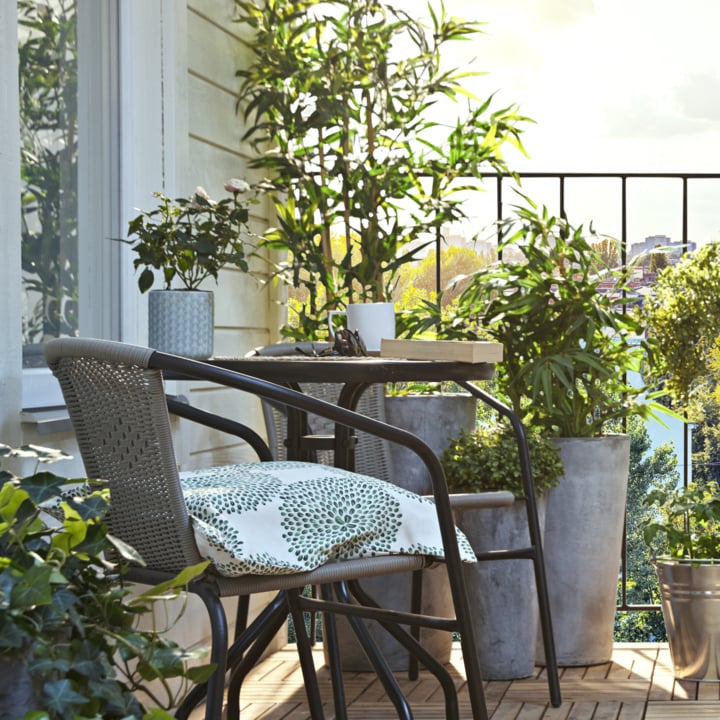 Gemütlicher Balkon mit Holzboden, Tisch, zwei gepolsterten Stühlen, Topfpflanzen, einer Tasse und Sonnenlicht, das durch das Grün fällt.