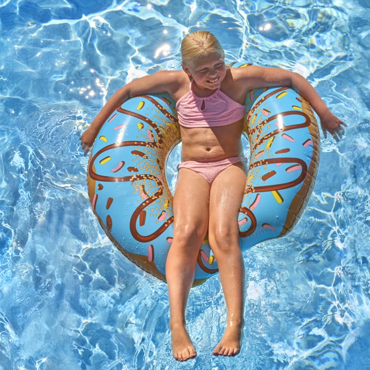Smilende jente i rosa badedrakt flyter på en blå donut-float dekket med dryssel, beina slepende i det solfylte svømmebassenget.