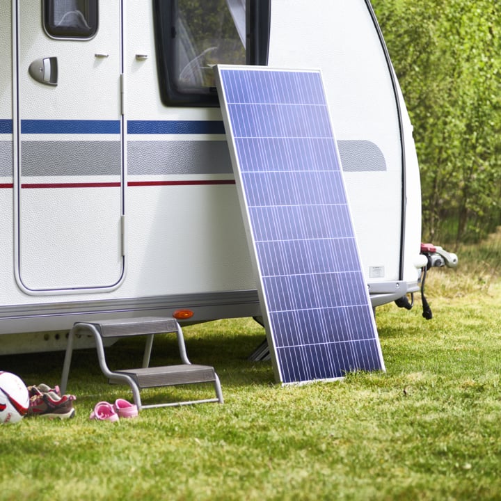Ein Solarpanel lehnt an einem Wohnwagen auf dem Gras, mit einem Metallhocker, einem Fußball und pinken Kinderschuhen in der Nähe mitten im Grün.