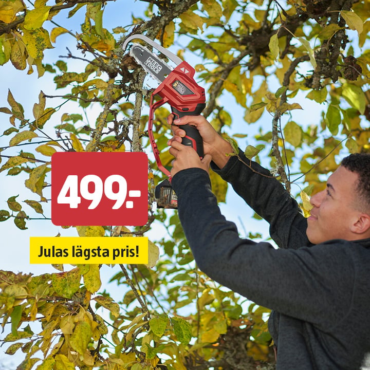 En person beskär trädgrenar utomhus med en sladdlös elsåg. På en röd skylt står priset 499:-, och på en gul etikett nedanför står det ”Julas lägsta pris!”. I bakgrunden syns gröna och gula löv.