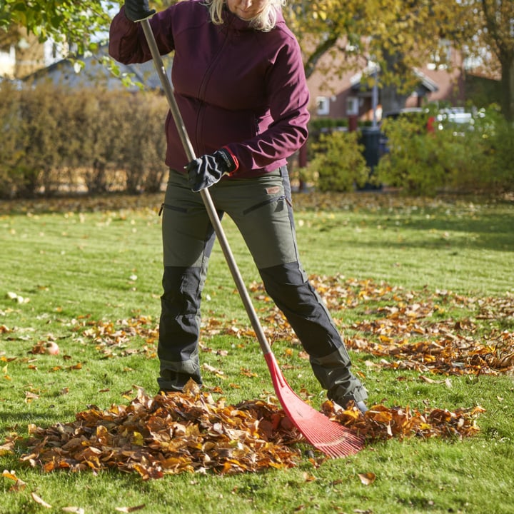Mit Handschuhen, einer kastanienbraunen Jacke und grünen Hosen harkt eine Person gefallene Blätter zu einem Haufen auf dem Gras. Im Hintergrund sind Bäume mit bunten Herbstblättern und mehrere Häuser zu sehen.
