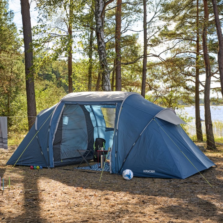 Ein geräumiges blaues Zelt steht auf mit Kiefernnadeln übersätem Boden zwischen Bäumen an einem See, umgeben von Klappstühlen, einem Tisch, Bällen und einer Matte. Sonnenlicht strömt unter klarem Himmel durch die Äste.