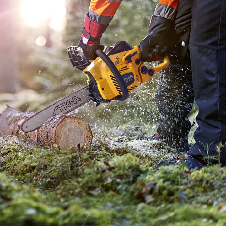 Jemand mit Handschuhen und Sicherheitsausrüstung bedient eine gelbe Stiga-Kettensäge, um draußen einen Baumstamm zu sägen, während Holzspäne verstreut und Sonnenlicht durch die Bäume dahinter strömt.