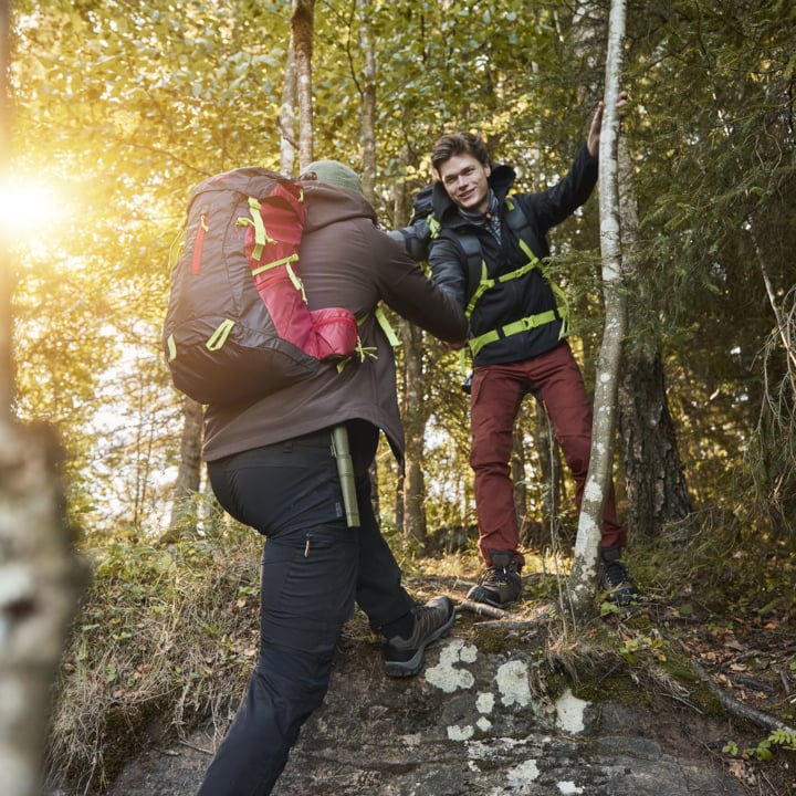 Zwei Wanderer in Outdoor-Kleidung machen sich auf den Weg einen felsigen Hang in einem Wald. Einer unterstützt den anderen, indem er seinen Arm hält, während Sonnenstrahlen durch die Bäume hinter ihnen scheinen.